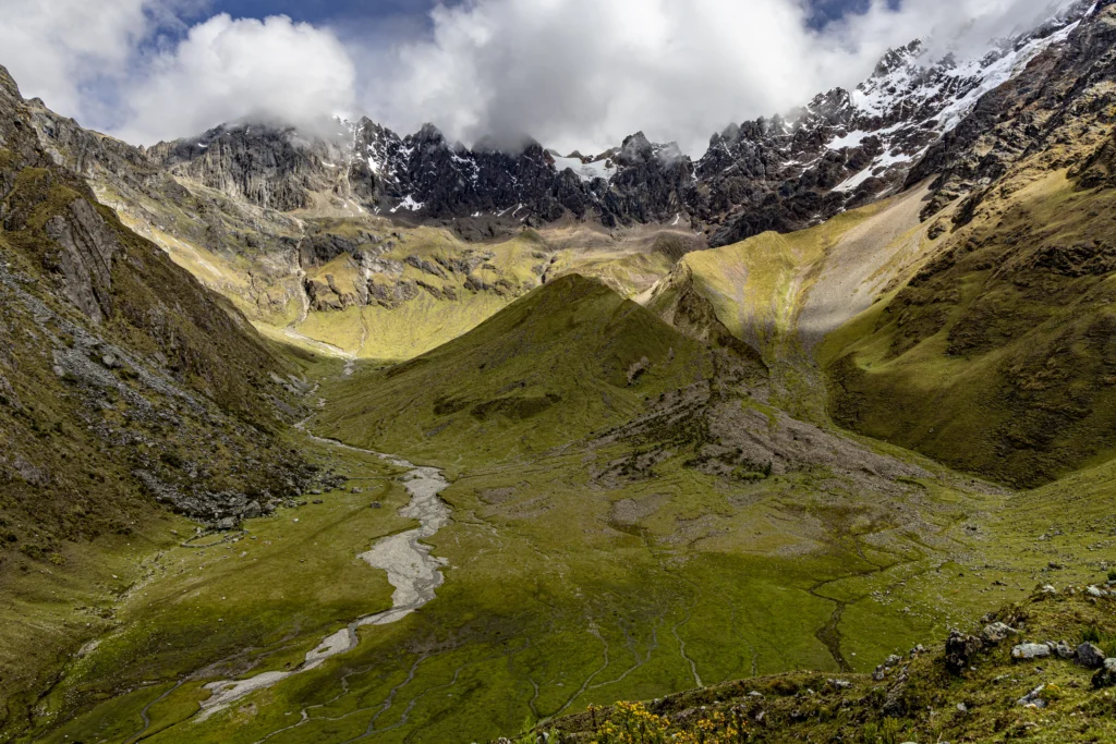 Panoramic alpine valley with a winding stream, grassy slopes, and rugged snow-capped mountains under cloudy skies for a dramatic landscape.