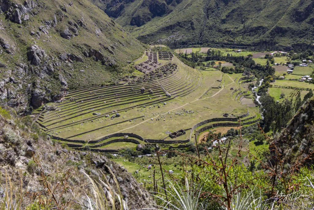 Aerial view of Patallacta's stepped stone terraces carved into a hillside with a valley and small village in the distance.