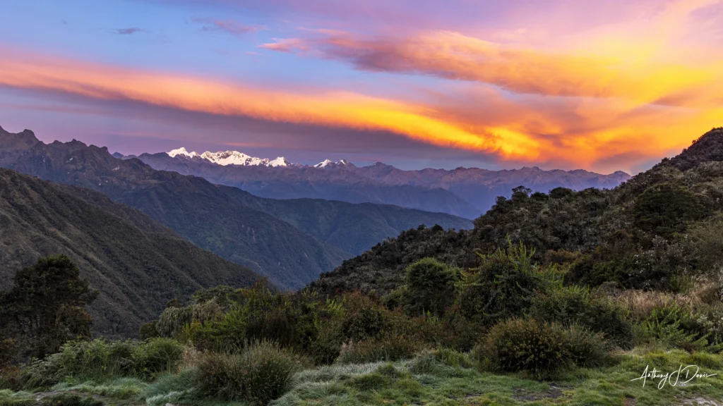 Sunset over the Vilcabamba mountain range with orange, pink, and purple sky and green foreground shrubs and hillsides