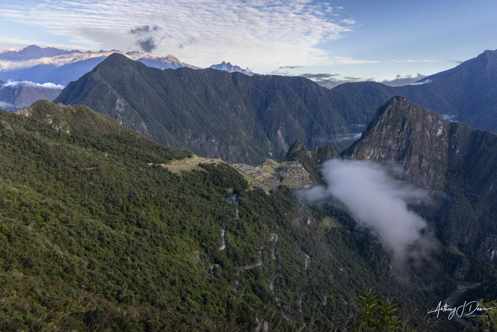 Panoramic view of Machu Picchu in front of steep green mountains with terraced fields in the valley and wispy clouds around the peaks.