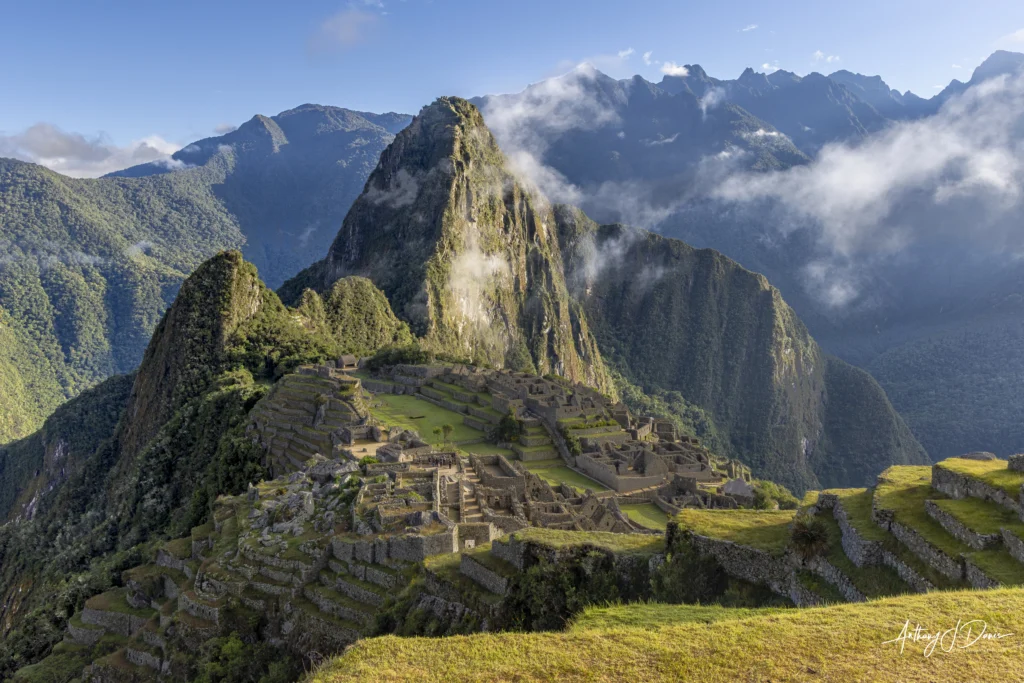 Ancient Machu Picchu stone terraces descend a hillside with green mountains and wisps of clouds in the background.