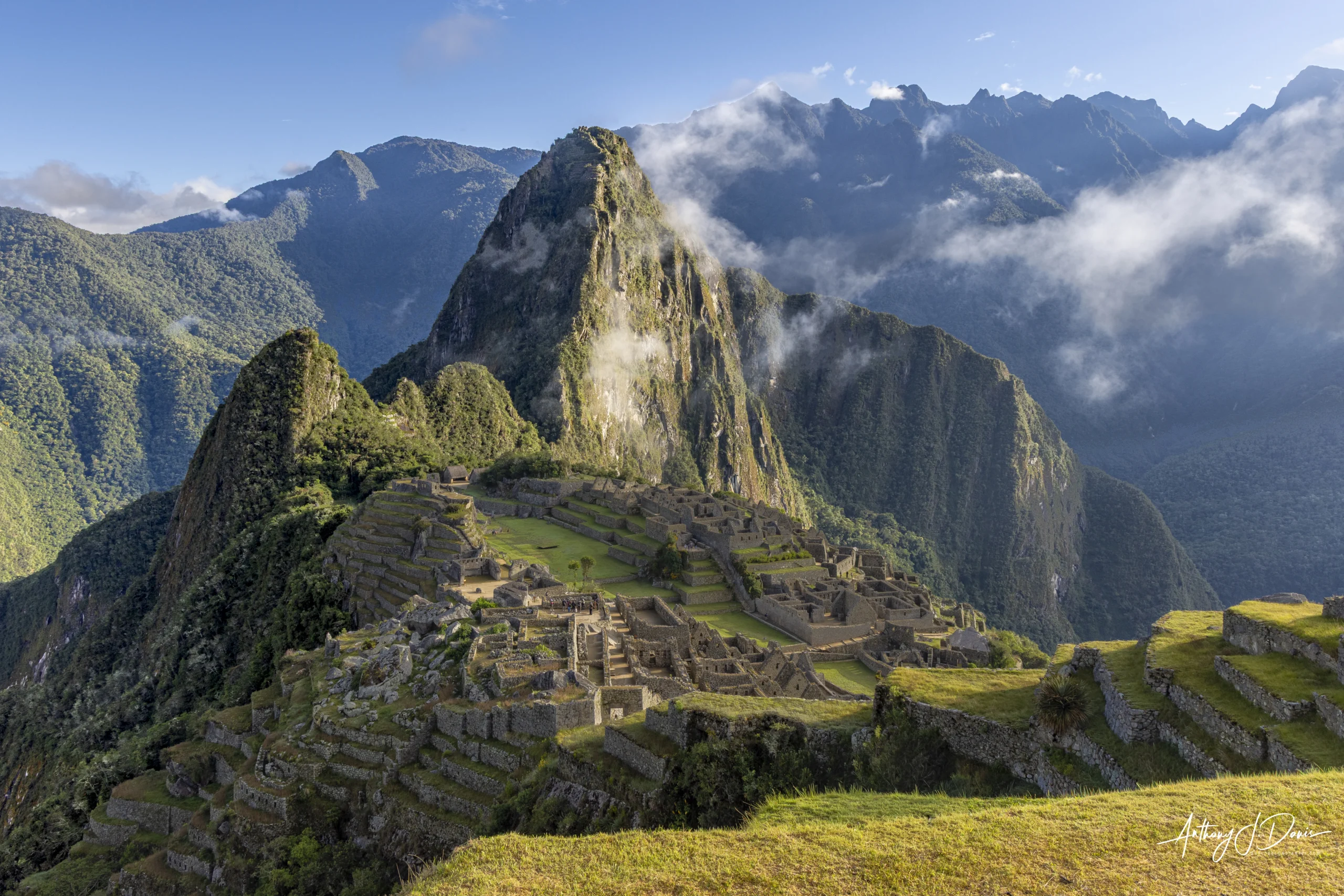 Ancient Machu Picchu stone terraces descend a hillside with green mountains and wisps of clouds in the background.