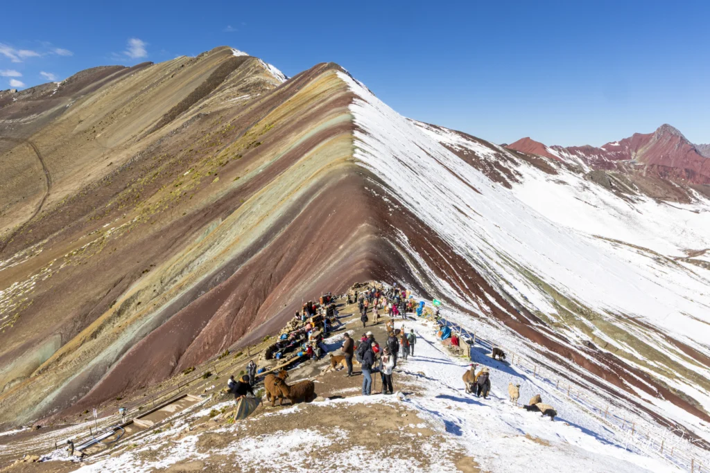 Colorful striped mountain ridge with a line of hikers and alpacas on a snow-covered slope under a blue sky.
