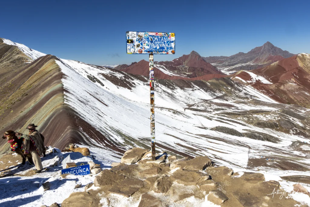 Two hikers stand near a sticker-covered sign at Rainbow Mountain (Vinicunca) with snow and colorful, striped peaks in the background.