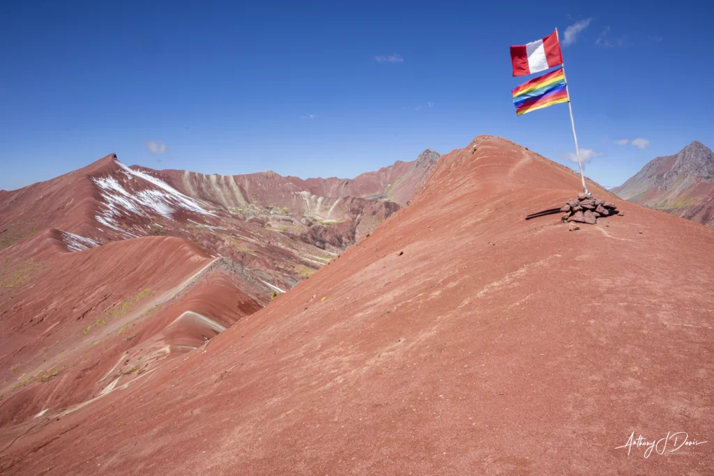 Wide view of red desert hills under a bright blue sky, with a tall flagpole and rainbow flag on a rocky summit.
