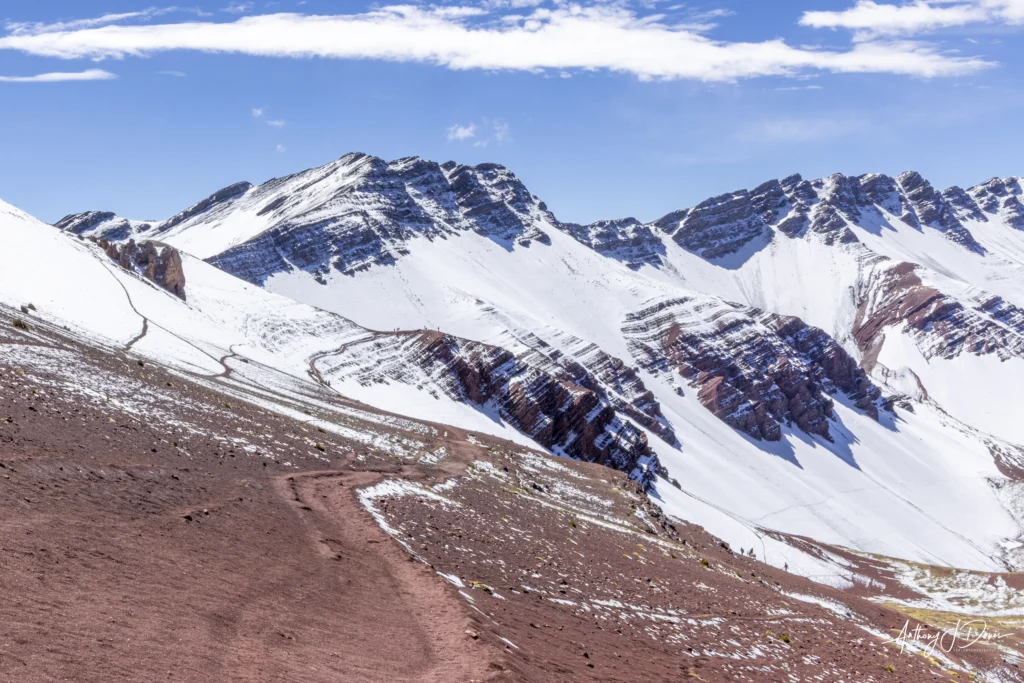 Snow-covered mountain ridge under a bright blue sky, with a reddish-brown rocky foreground and a few hikers in the lower right corner.